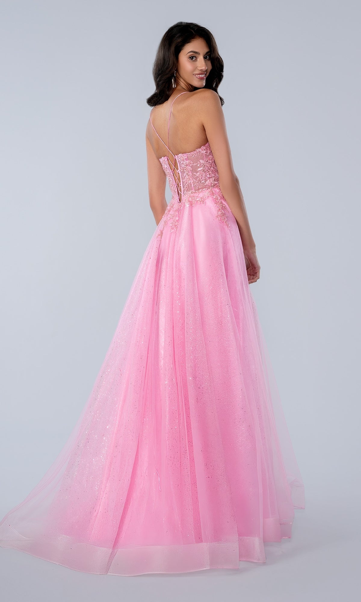 A woman faces away, wearing the Stella Couture 25140 Glitter Prom Ball Gown—long, light pink with a v-neck corset bodice, delicate lace details, and thin crisscross straps against a plain light background.